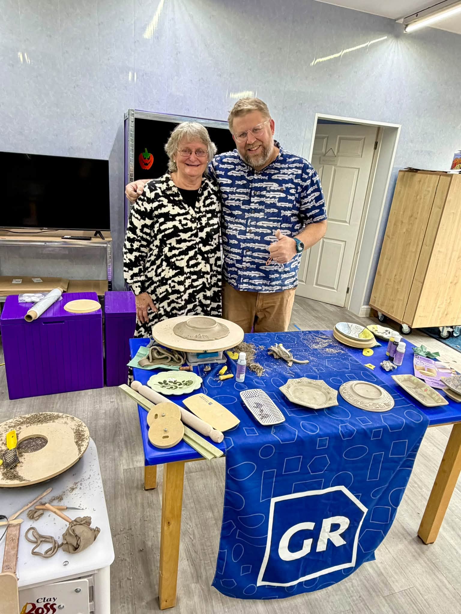 Jeff Rottman with a workshop attendee beside finished clay pieces and GR Pottery Forms